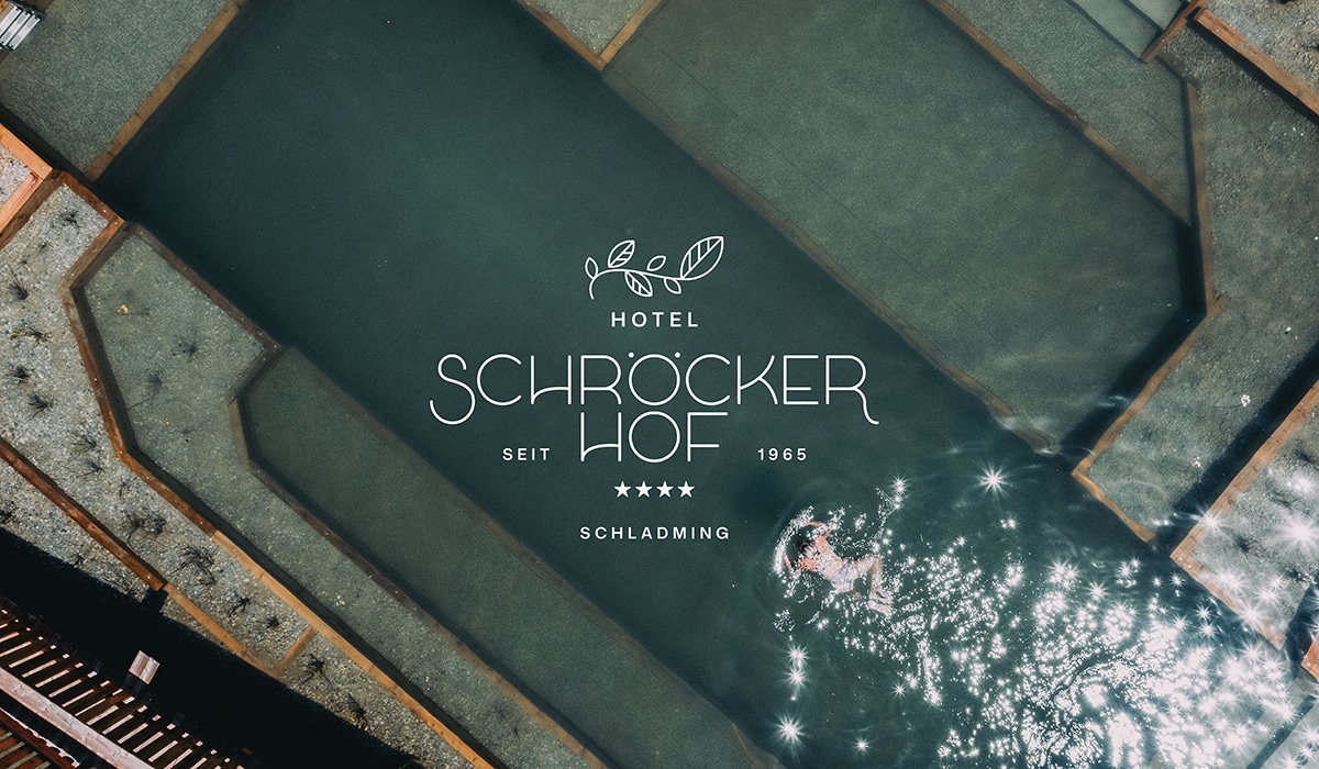 Luftaufnahme eines modernen Naturpools mit schwimmender Person im Sonnenlicht, überlagert vom Logo des 4-Sterne-Hotels Schröckerhof in Schladming.